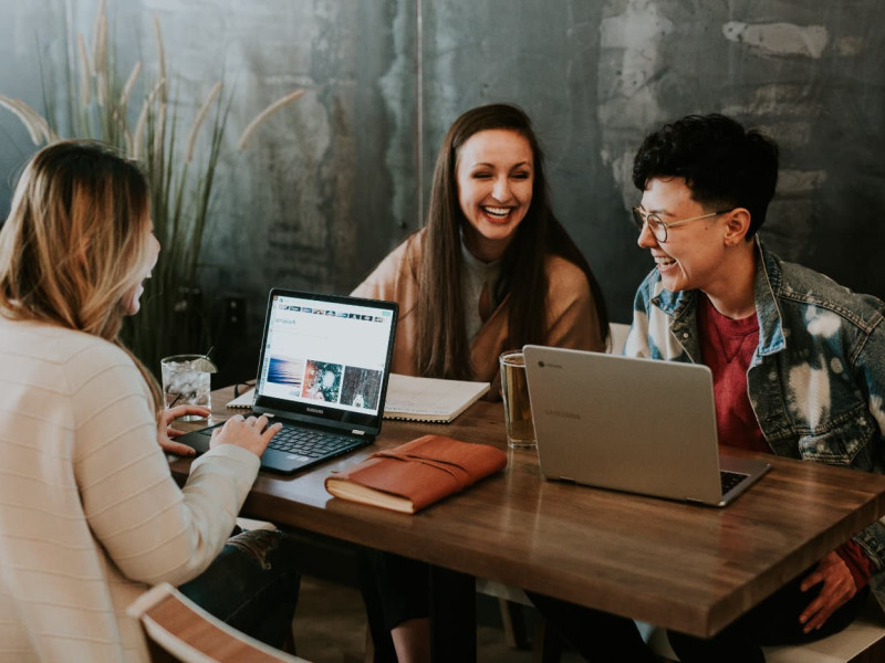 Three women sit at a desk with their laptops. They are chatting and laughing.