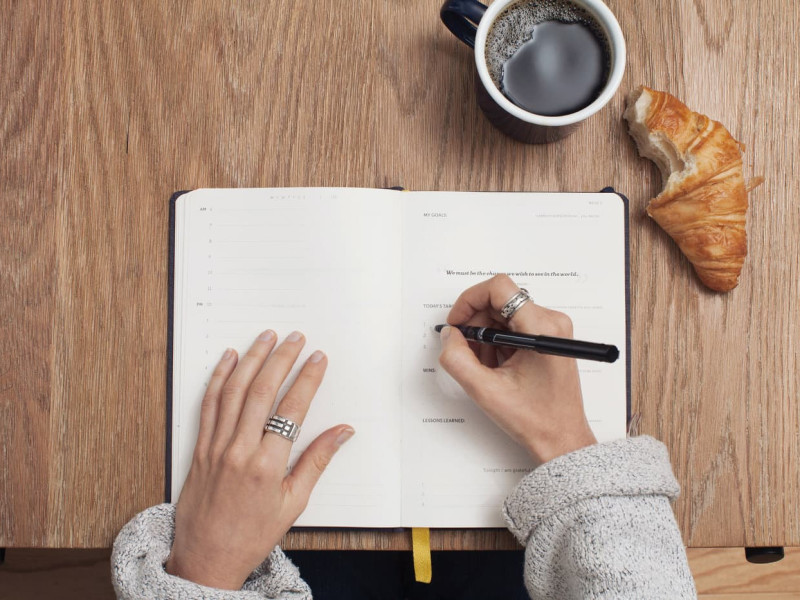 A table with a notebook, a coffee mug and a croissant from above. A women is taking notes.